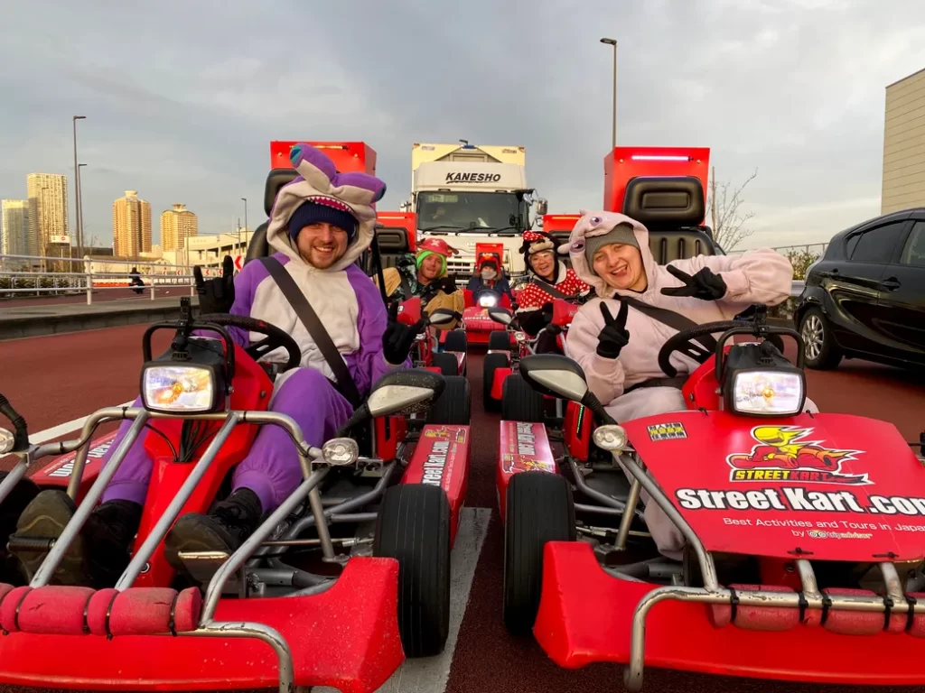 IMG_0829 - StreetKart Group of people in red go-karts wearing colorful animal costumes, posing and waving on a track with a city skyline in the background.