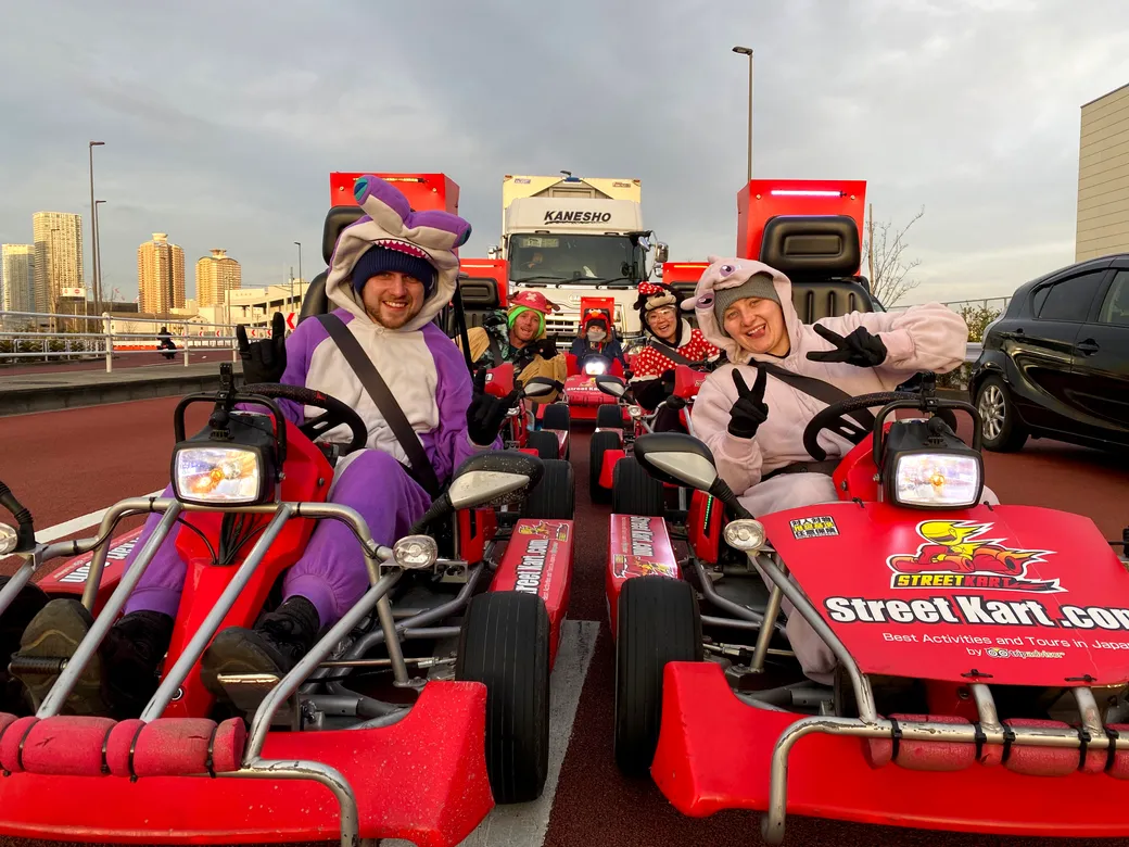Group of people in red go-karts wearing colorful animal costumes, posing and waving on a track with a city skyline in the background.