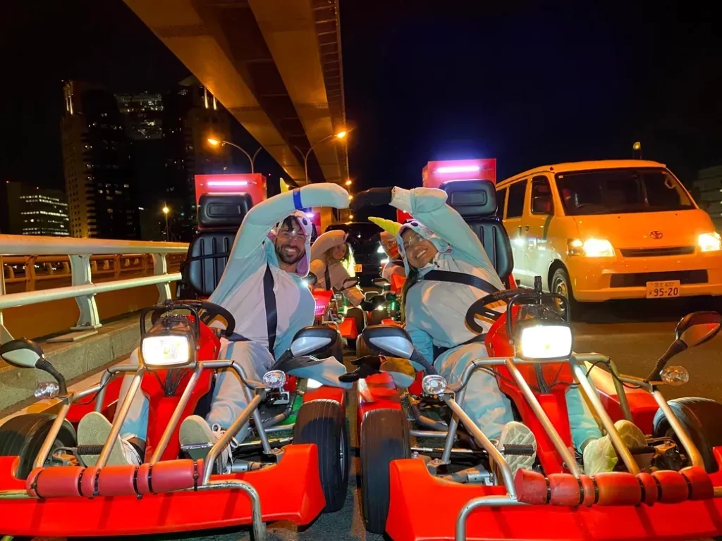 Two people in white costumes drive red go-karts on a nighttime city bridge, forming a heart shape with their arms.