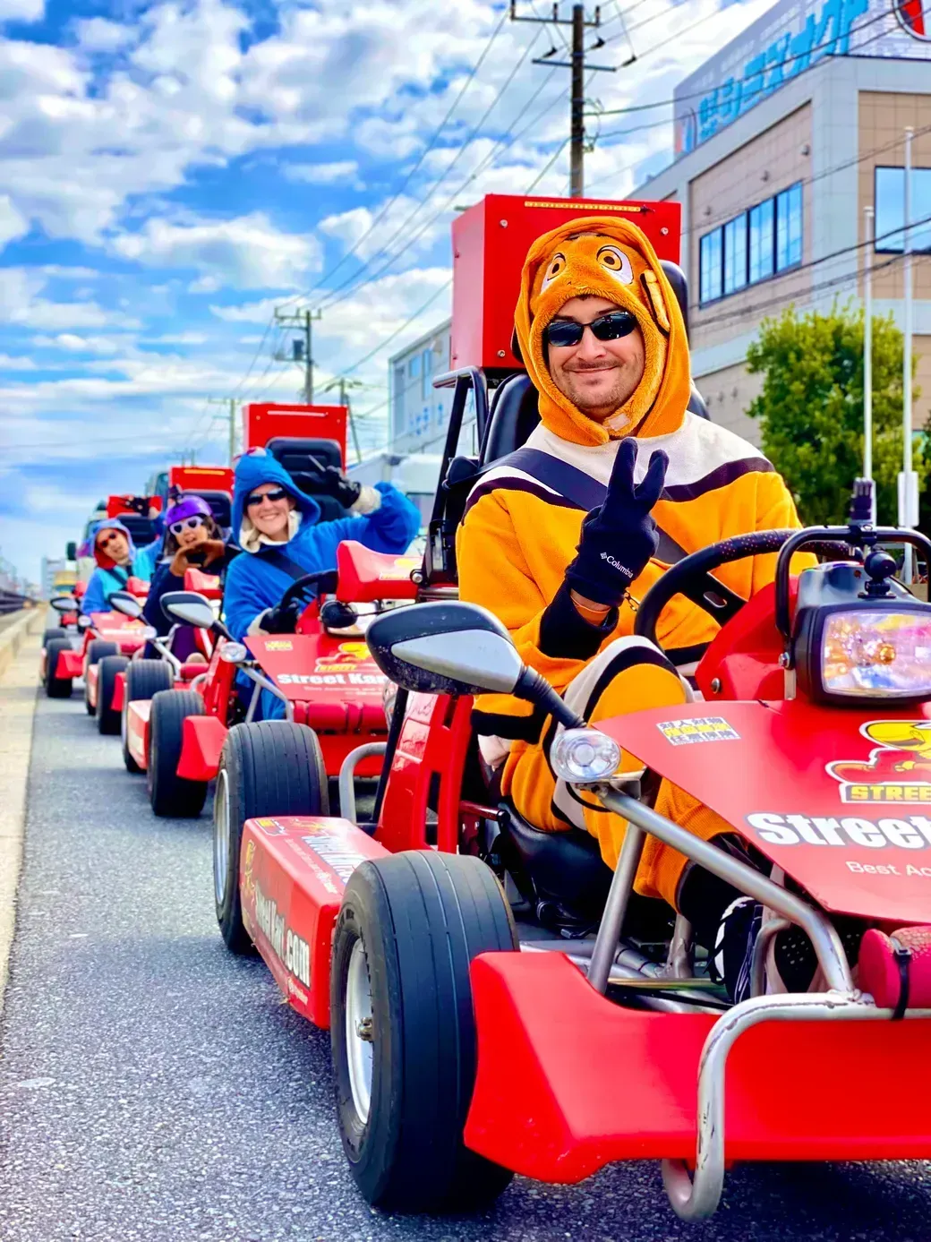 Man in an orange animal onesie driving a red go-kart, wearing sunglasses and flashing a peace sign