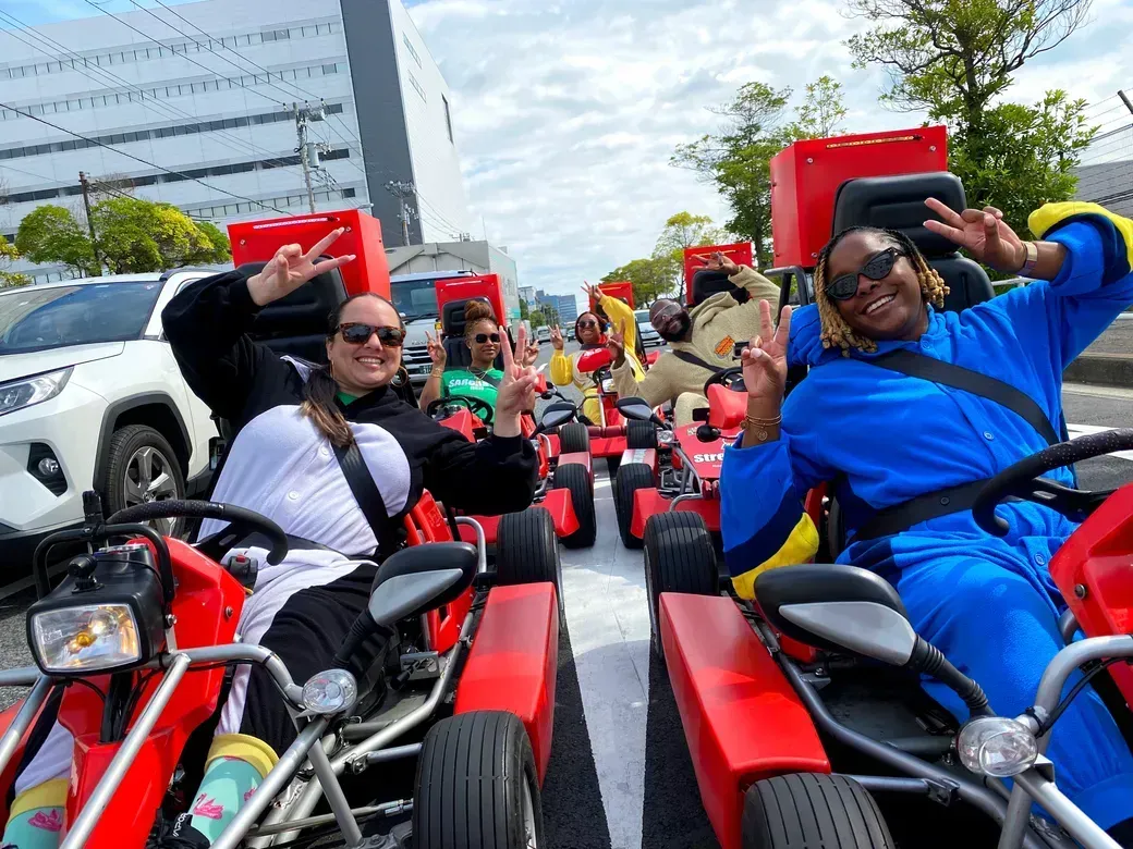 Group of people riding red pedal go-karts on a city street, waving peace signs and smiling at the camera.